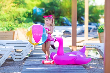 Baby girl on beach with pink flamingo dressed in stylish swimming wear posing on wooden floor. Scene on vacation on sea side of a cute lady smiling and enjoy life time childhood infancyの写真素材