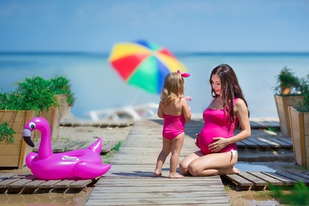 Pregnant woman on beach healthy vacation posing with cute baby daughter wearing pink stylish swimming wear. Family love scene of beautiful lady and funny child girlの写真素材