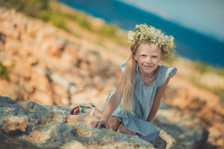 Blond girl pretty with blue eyes wearing light dress with arms and legs and wild flowerrs wreath on top head posing in ancient city stones close to seaside beach with teddy bear.の写真素材