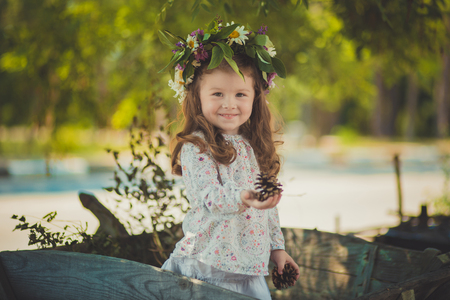 Cute young girl with brown eyes brunnette plait hair and pink cheek wearing white dress shirt and posing close to wooden house fence wagon looking to camera with wreath of flowers on head.の写真素材