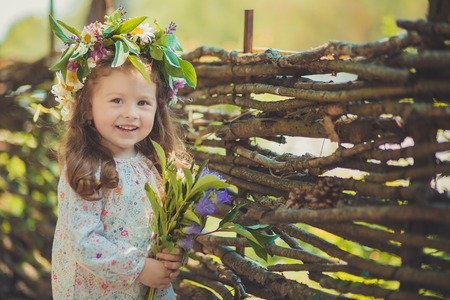Cute young girl with brown eyes brunnette plait hair and pink cheek wearing white dress shirt and posing close to wooden house fence wagon looking to camera with wreath of flowers on head.の写真素材