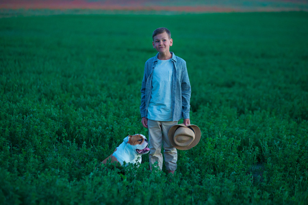 Little boy sitting standing with his english bull dog on the meadow of green rye. Handsom kid posing with best friend wearing stylish clothes and rustic retro hat.の写真素材