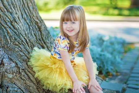 Little girl having fun in city park close to tree and fountain wearing stylish yellow dress posing and smiling on camera.の写真素材
