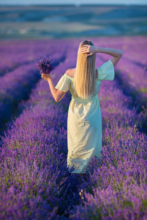 Smiling beautiful blond lady model on lavender field enjoy summer day wearing airy whit dress with bouquet of flowersの写真素材