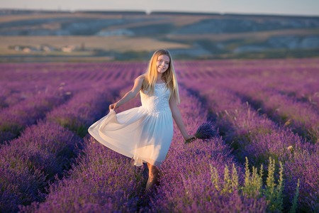 Smiling beautiful blond lady model on lavender field enjoy summer day wearing airy whit dress with bouquet of flowersの写真素材