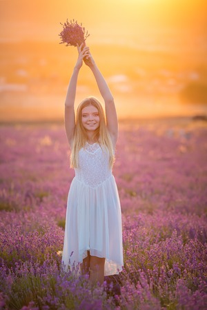 Smiling beautiful blond lady model on lavender field enjoy summer day wearing airy whit dress with bouquet of flowersの写真素材