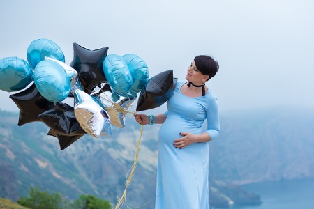 Pregnant woman posing on mountain coast line wearing blue long dress with air baloonsの写真素材