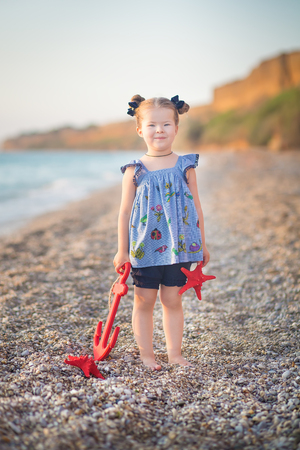 Cute little girl enjoying summer time on sea side beach happy playing with red star and tiny toy anchor sand wearing nobby clothes with brunette hairs.の写真素材