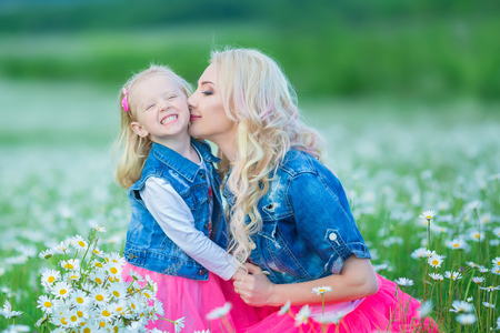 Mom and daughter on a picnic in the chamomile field. Two beautiful blondes wearing jeans jacket and pink skirt. Mother and baby girl embracing in the chamomile field with basket of bouquet dandalion.の写真素材