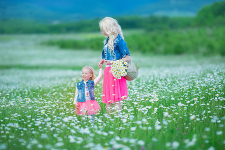 Mom and daughter on a picnic in the chamomile field. Two beautiful blondes wearing jeans jacket and pink skirt. Mother and baby girl embracing in the chamomile field with basket of bouquet dandelion.の写真素材