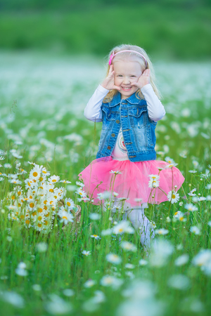 Cute little smiling baby girl in chamomile field. Little blond child with wreath on head in chamomiles wearing jeans and pink skirt enjoying hide-and-seek, whoop, peek a boo.の写真素材