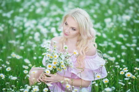 Attractive blonde in chamomile field. Young woman in wreath surrounded by chamomiles wearing stylish pink dress enjoying summer spring.の写真素材