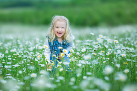 Cute little smiling baby girl in chamomile field. Little blond child with wreath on head in chamomiles wearing jeans and pink skirt enjoying hide-and-seek, whoop, peek a boo.の写真素材