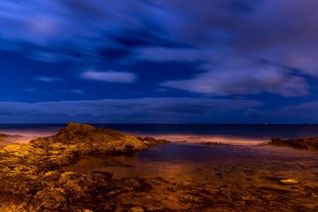 Long exposure shot of a cliff full of stones and a lagoon beside the ocean during a deep night as water poured onto the stones and the surrounding area.の写真素材