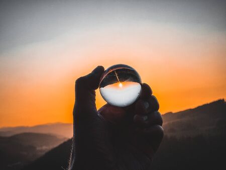 Taking photos of sunset through the small glass ball holding in handの写真素材