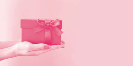 Woman's hands holding red gift box with bow on pink background. Side view, holiday present concept with copy space, banner.の写真素材
