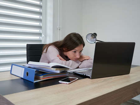 A young woman works at home and looks at the laptop monitor. Tired Girl sits at the table and props her chin in her arms. There are binders with documents on the table. Home officeの写真素材