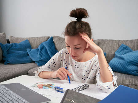 Young worried woman doing financial balance. The girl is counting the bills. There is money in euros and a laptop on the table. Summary of monthly costs. Rising living costs cause stressの写真素材