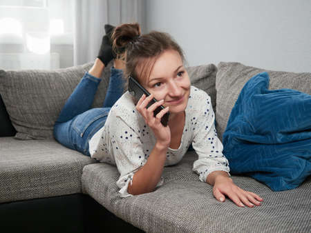 A young woman is lying on the couch in the living room and is using a smartphone. A relaxed and smiling girl talking on the phone. Video call.の写真素材