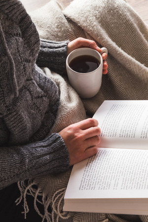 Woman wearing gray woolly cardigan enjoying a mug of tea and reading a book under a cozy blanket, hygge conceptの写真素材