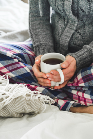 Woman wearing cozy pyjamas and gray cardigan drinking tea on a bedの写真素材