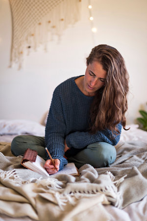 Woman in blue sweater journaling in leatherbound notebook whilst sat on bedの写真素材