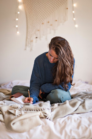 Woman in blue sweater sat on bed writing in her leatherbound journal with a pencilの写真素材