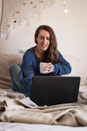 Woman in blue sweater sat on bed using laptop computer and drinking mug of tea or coffee