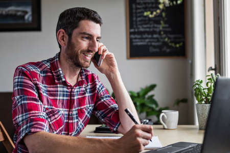 Smiling businessman on phone call in home office, writing notes in front of laptop