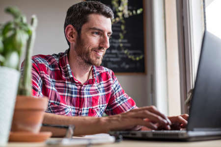 Smiling man sat at desk writing on laptop at homeの写真素材