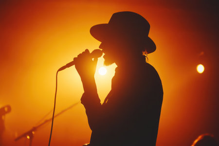 Artist with hat singing on stage under warm orange backlight, representing emotion, music, and creative self-expression.の素材