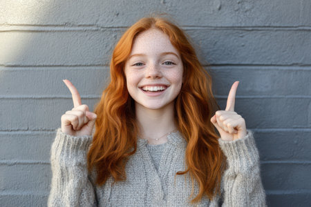 Smiling young ginger teenage girl pointing upwards towards copy space, radiating excitement, joy, and vibrant youthful energy.の素材