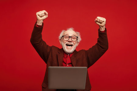 Elderly man with glasses joyfully celebrates achievement in front of a laptop, symbolizing triumph, technology use, and happiness.の素材