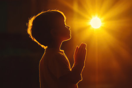 Silhouette of a child praying to God under glowing sunset light, symbolizing faith, hope, spirituality, and deep religious belief.の素材