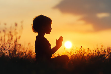 Silhouette of a child praying to God under glowing sunset light, symbolizing faith, hope, spirituality, and deep religious belief.の素材