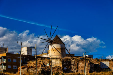 A rustic windmill stands amid old brick buildings under a bright blue sky with fluffy cloudsの写真素材