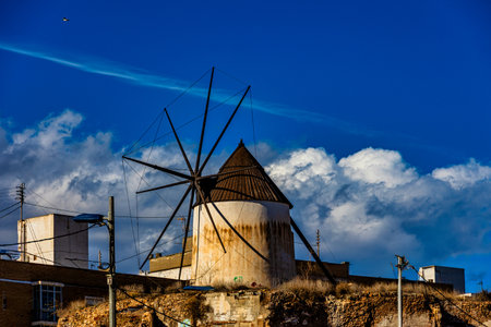 A rustic windmill stands amid old brick buildings under a bright blue sky with fluffy cloudsの写真素材