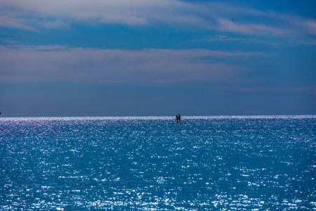 Sunlight shimmers across a vast expansion of calm water. Two silhouettes, possibly paddleboarders,の写真素材