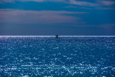 Sunlight shimmers across a vast expansion of calm water. Two silhouettes, possibly paddleboarders,の写真素材