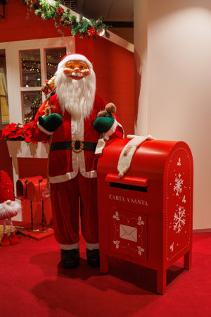 Santa Claus stands next to a red mailbox designated for letters to him, surrounded by festive decorations.の写真素材