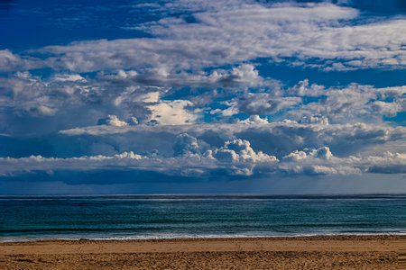 Fluffy white clouds stretch across a vast blue sky above a serene ocean horizon, creating a tranquil scene. The reflection of the clouds on the water adds to the peaceful ambiance.の写真素材