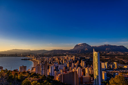 A coastal cityscape is bathed in the warm glow of a setting sun, with high-rise buildings clustering along the shorelineの写真素材