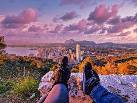 Two people sit on a rocky ledge overlooking a cityscape with a vast view of buildings, mountains, and coastline under a clear blue sky.の写真素材