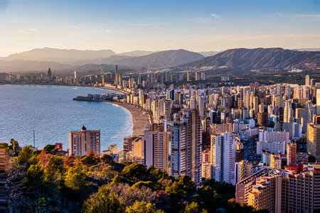 A coastal cityscape Benidorm is bathed in the warm glow of a setting sun, with high-rise buildings clustering along the shorelineの写真素材