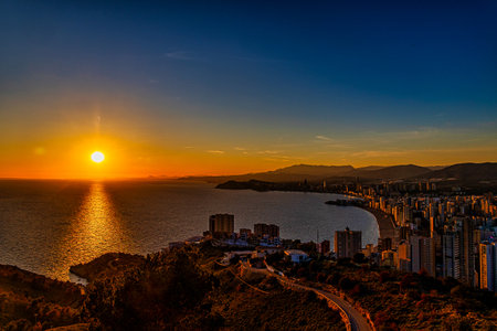 A stunning sunset casts a warm glow over a coastal city, with the sun reflecting beautifully on the calm seaの写真素材