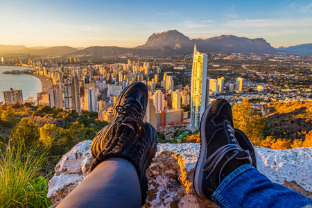 Two people sit on a rocky ledge overlooking a cityscape with a vast view of buildings, mountains, and coastline under a clear blue sky.の写真素材