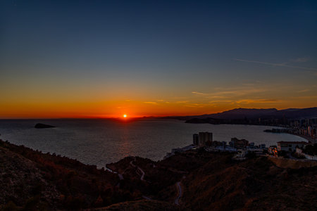 A stunning sunset casts a warm glow over a coastal city, with the sun reflecting beautifully on the calm seaの写真素材