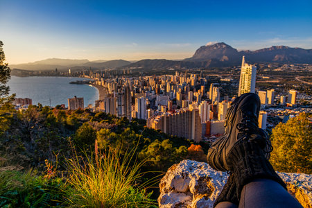 a person sits on a rocky ledge overlooking a cityscape with a vast view of buildings, mountains, and coastline under a clear blue sky.の写真素材