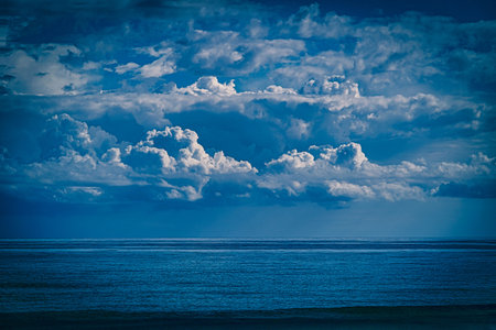 Fluffy white clouds stretch across a vast blue sky above a serene ocean horizon, creating a tranquil scene. The reflection of the clouds on the water adds to the peaceful ambiance.の写真素材