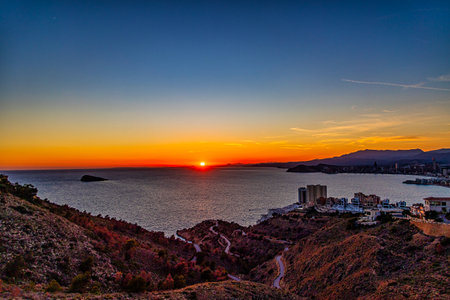A stunning sunset casts a warm glow over a coastal city, with the sun reflecting beautifully on the calm seaの写真素材